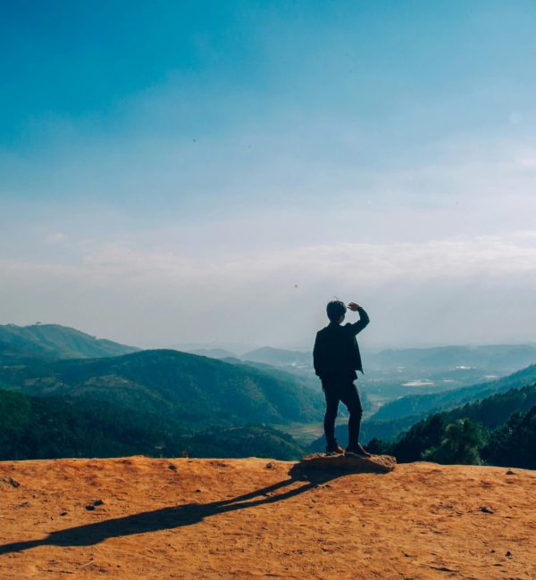 A lone traveler stands on a hilltop, gazing over a vast mountain landscape under a clear blue sky.